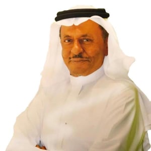 Man in traditional attire seated with arms crossed on a white background.