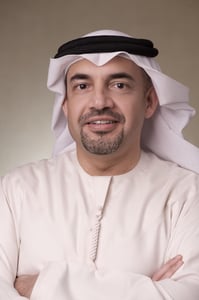 Man in white thobe and black keffiyeh smiling against a brown background.