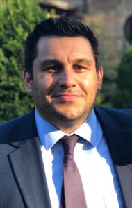 Man in a dark suit and light blue shirt with green foliage in the background.