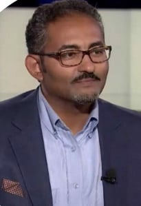 Man in a suit with glasses sitting in a television studio setting.