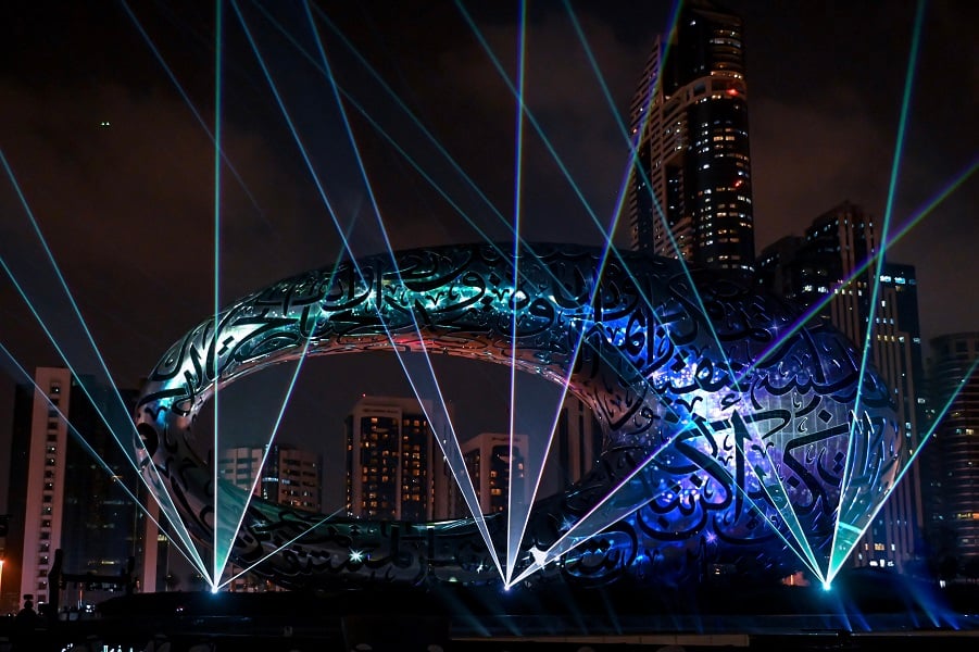 Calligraphic sculpture illuminated by laser beams in a cityscape.