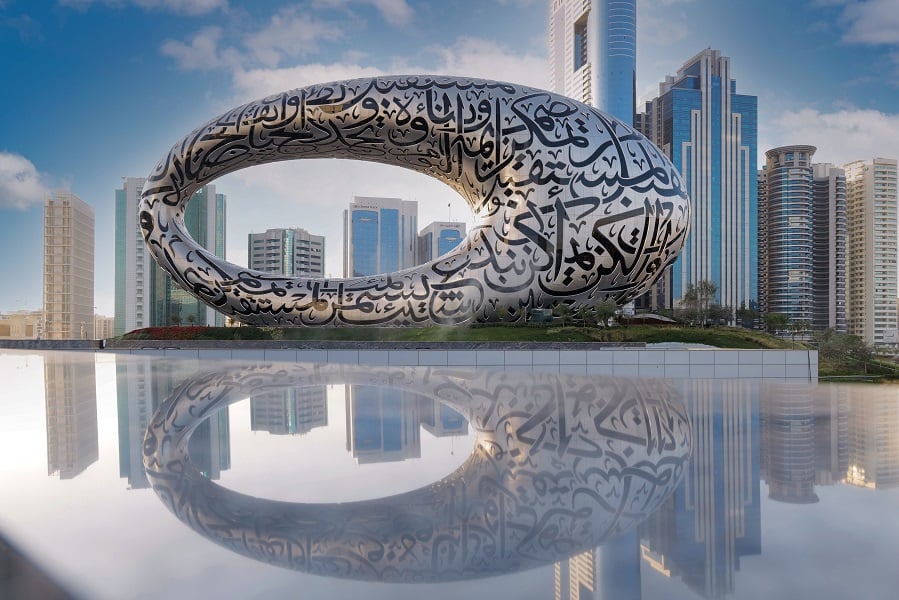 Calligraphic sculpture reflecting in water amidst skyscrapers under cloudy skies.