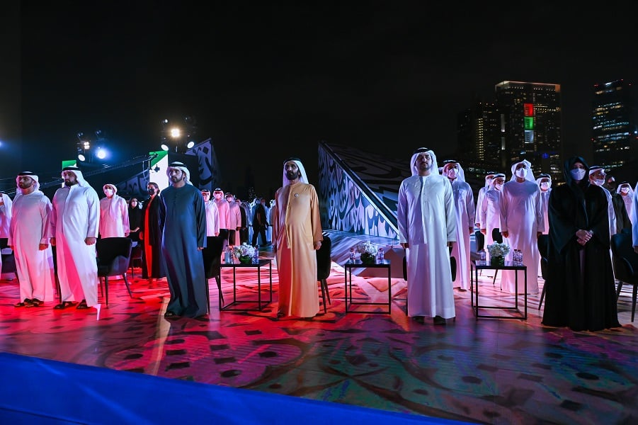 Men in Emirati attire attend outdoor event with modern cityscape backdrop.