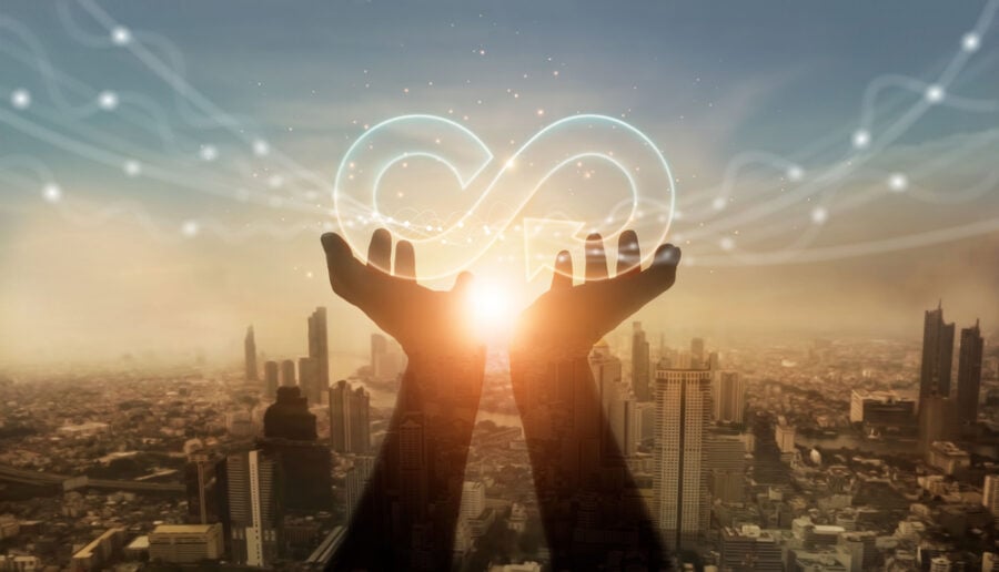 Hands holding glowing infinity symbol above city at sunset.