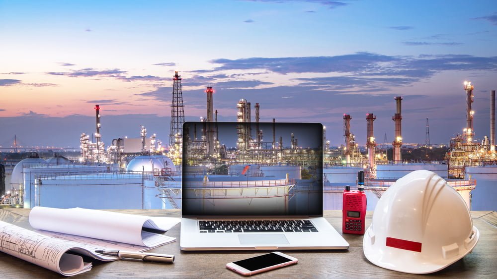 Laptop displays oil refinery with blueprints and hardhat on desk.