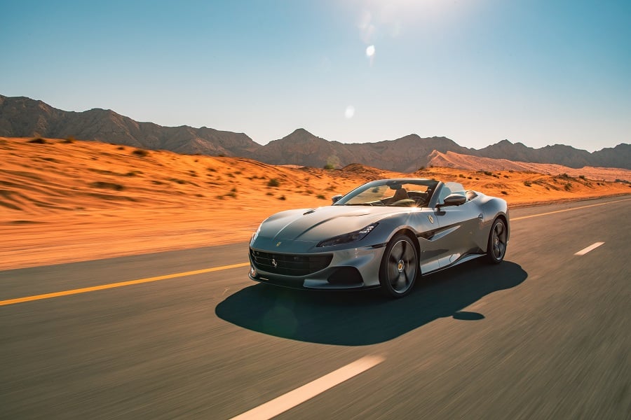 Silver Ferrari on a desert highway with dunes and mountains.