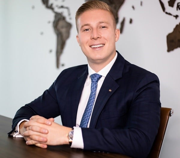 A young man in a suit smiles at the camera, sitting at a desk with a world map behind him.