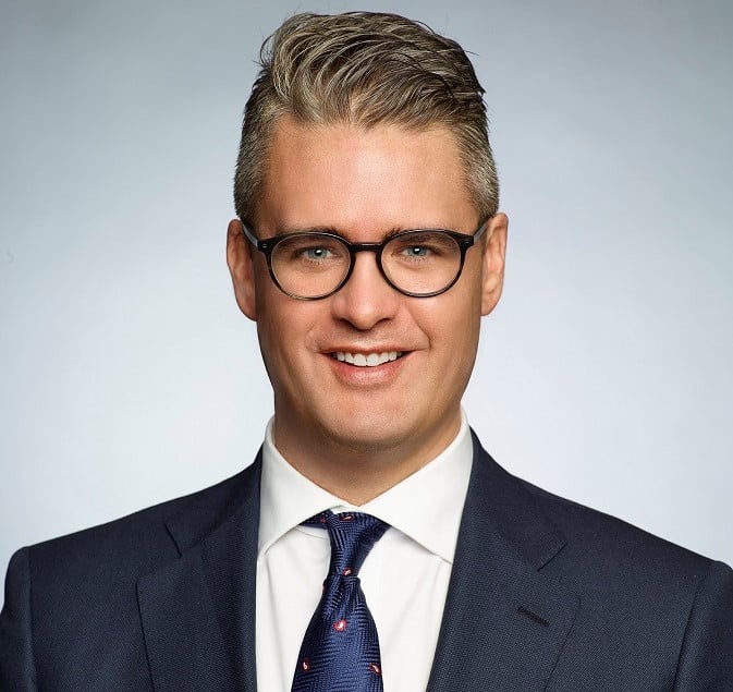 Man in a dark suit and glasses smiling with a gray background.