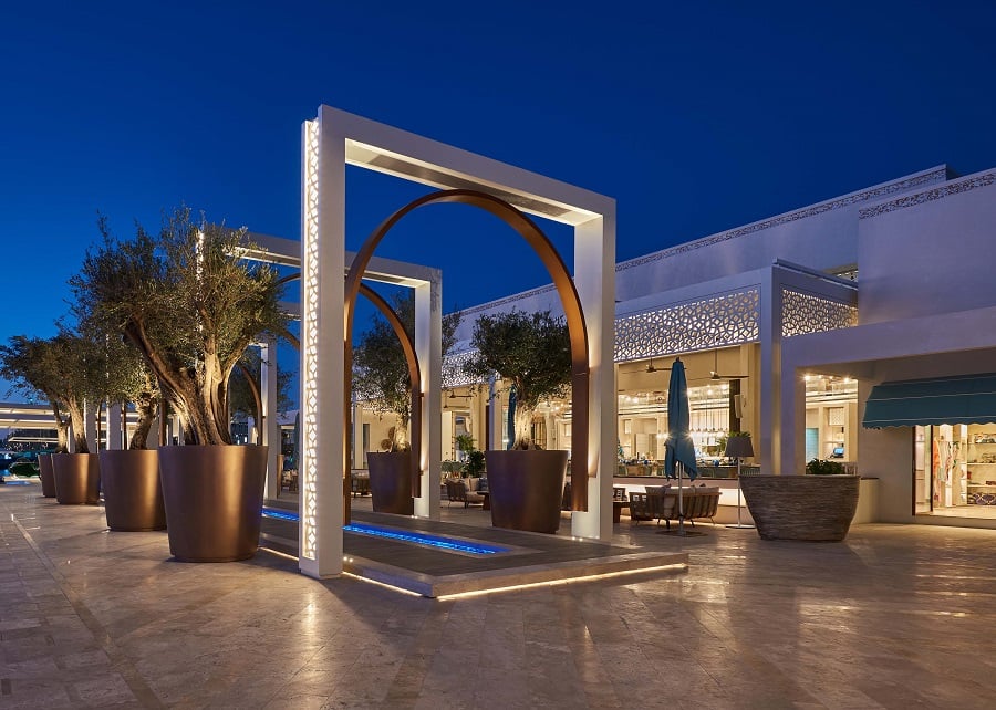 Modern archways illuminated at night frame olive trees leading to an outdoor restaurant.