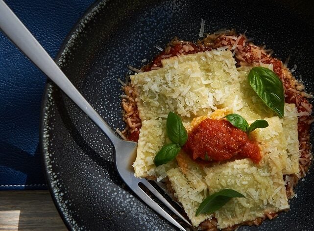 Ravioli topped with sauce, parmesan, and basil in a speckled bowl.