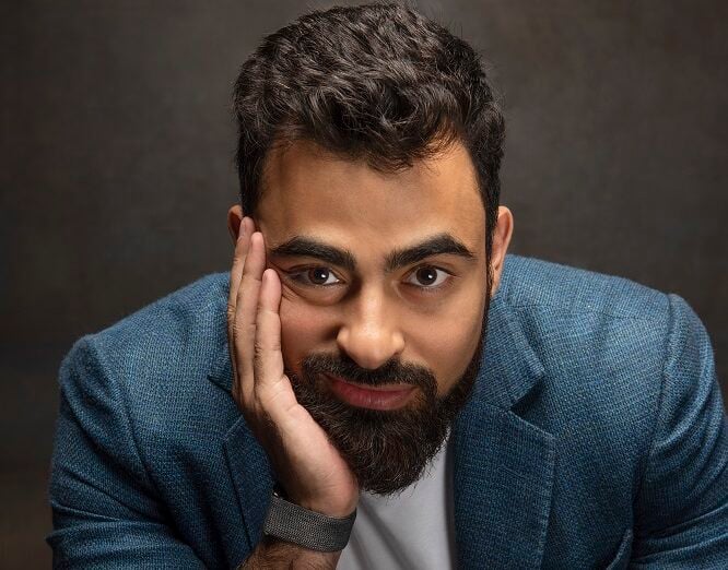 Bearded man in blue blazer looking pensive against dark background.