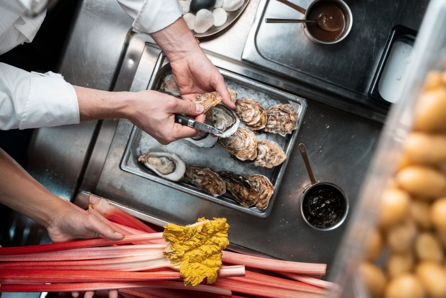 Chef shucking oysters on a metal tray with ingredients nearby.