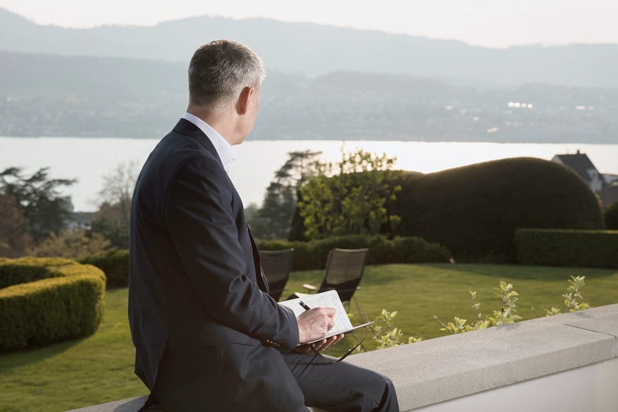 Man in suit writes by a lake and garden.