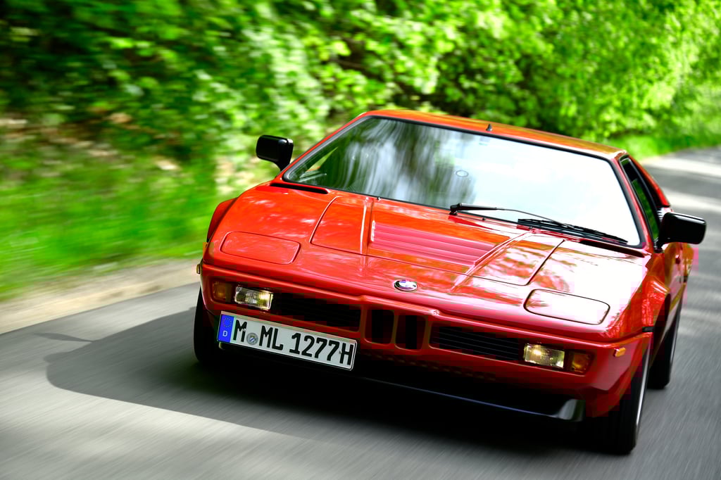 Red BMW M1 sports car on a country road with license plate.