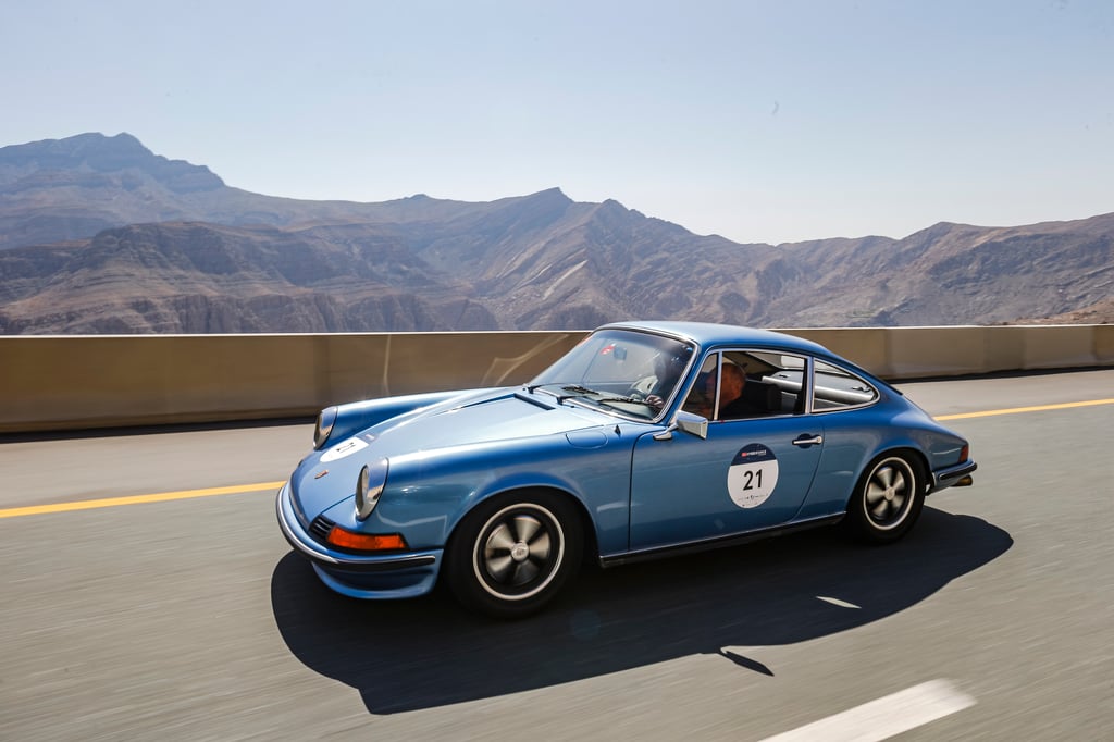 A light-blue Porsche 911 drives along a mountain highway with a visible