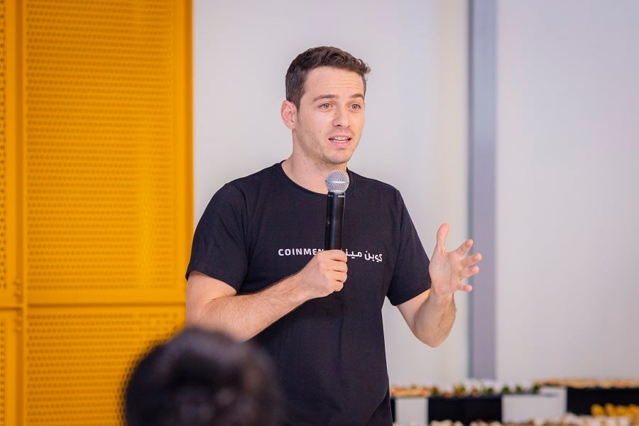 Man in COINMEN t-shirt speaking into a microphone with a yellow wall backdrop.