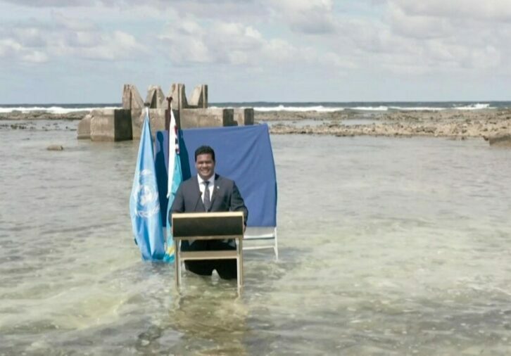 Man at podium in ocean water near submerged structures and flags.