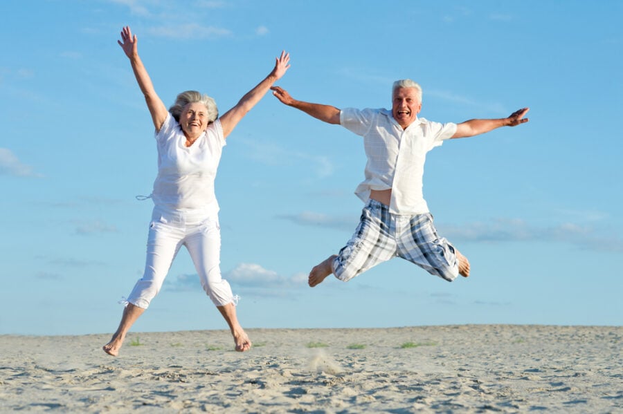 Two seniors jumping joyfully on a sandy beach under a blue sky.