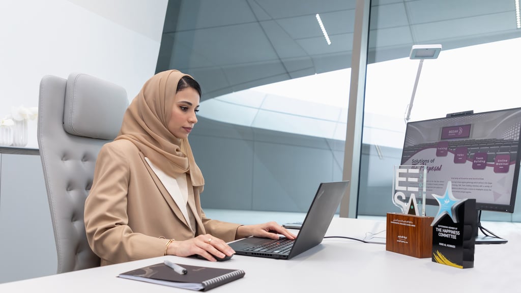 Woman in a hijab types at a desk with awards and a presentation visible in the office.