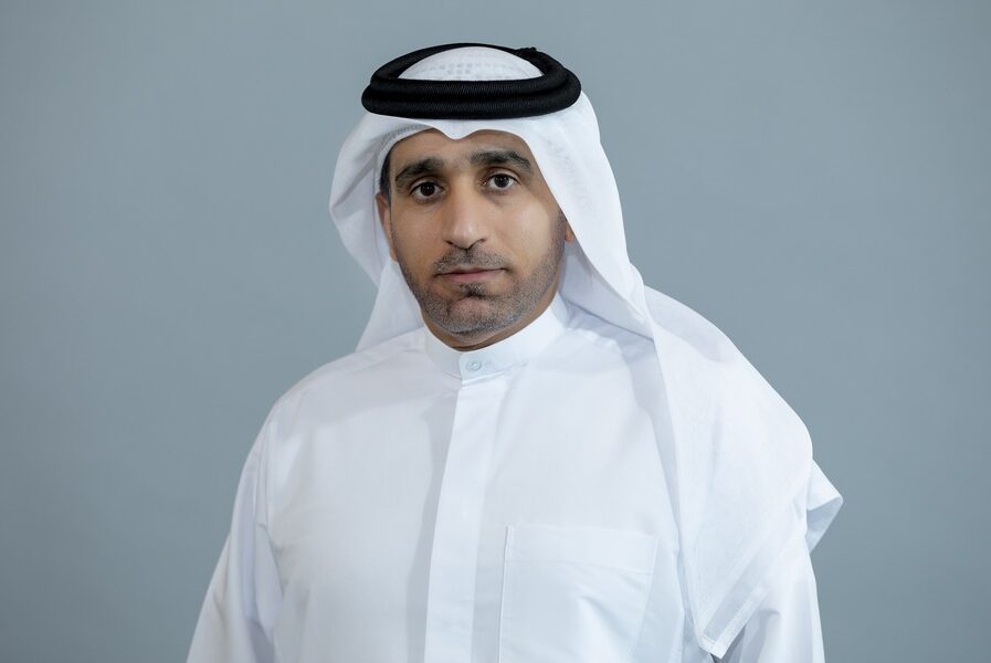 Man in white thobe and black head-ring against a grey background.