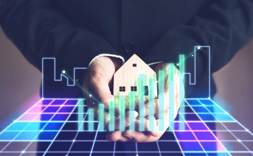 Hands hold a miniature house with an upward-trending real estate graph.
