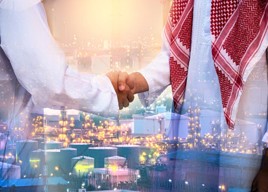 Two men shaking hands, one in Middle Eastern attire, with an industrial plant in the background.