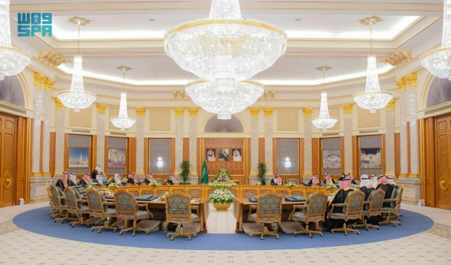 A group of people in formal attire sit around a large table in a palatial room.