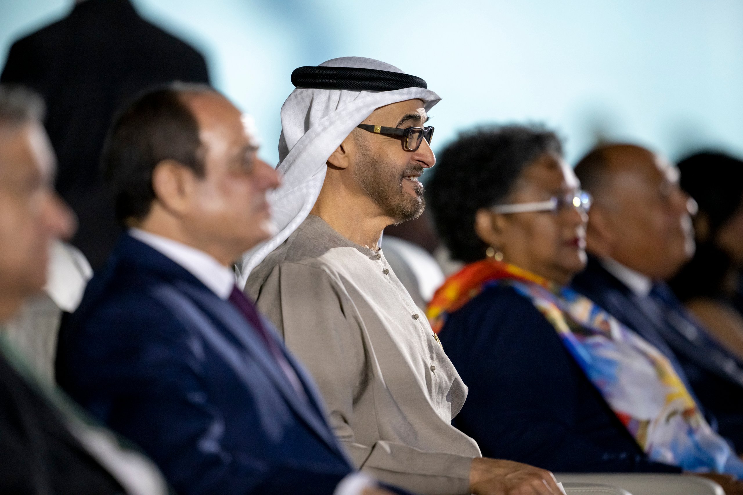 Man in kandura sits among attendees at outdoor event.