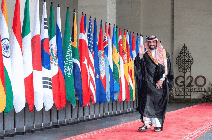 Man in Saudi attire waving on a red carpet at a G20 Indonesia event.