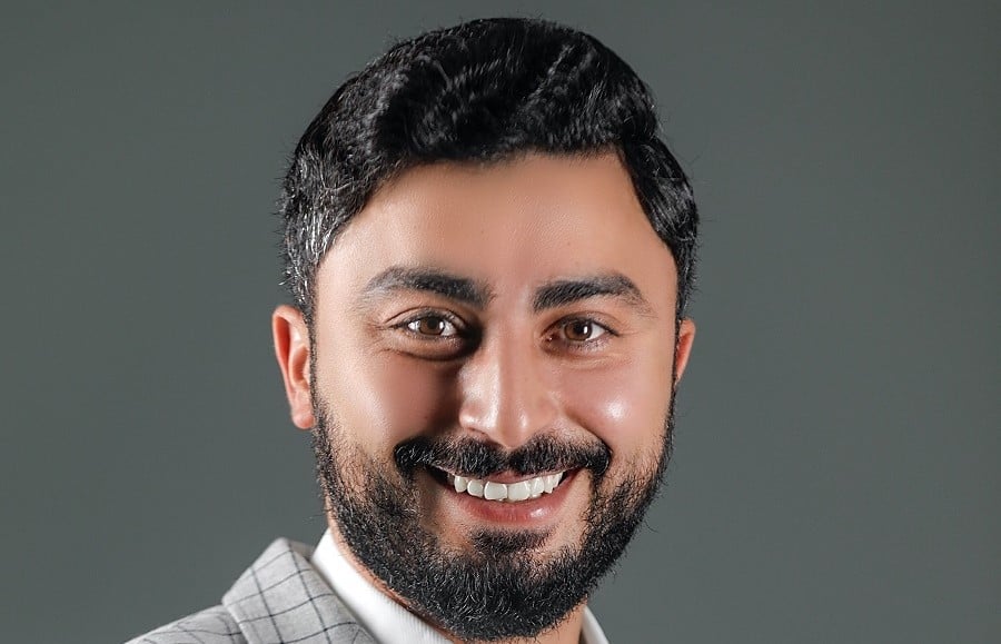Man with dark hair and beard smiling in a gray jacket.