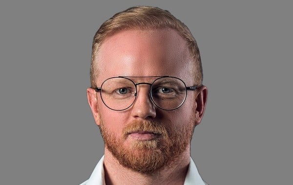 Man with red beard and glasses against gray background.