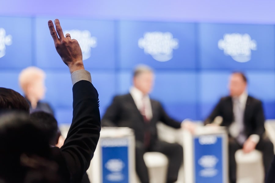 Person raising hand at panel discussion with blue logo backdrop.