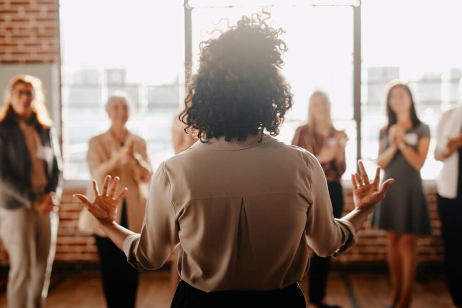 Woman addressing an applauding group of women in a sunlit room.