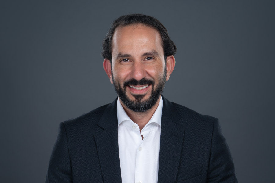 Man in a dark suit and white shirt smiling against a gray background.