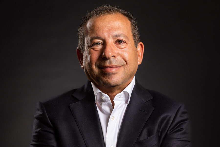 Middle-aged man in dark suit smiles against black background.