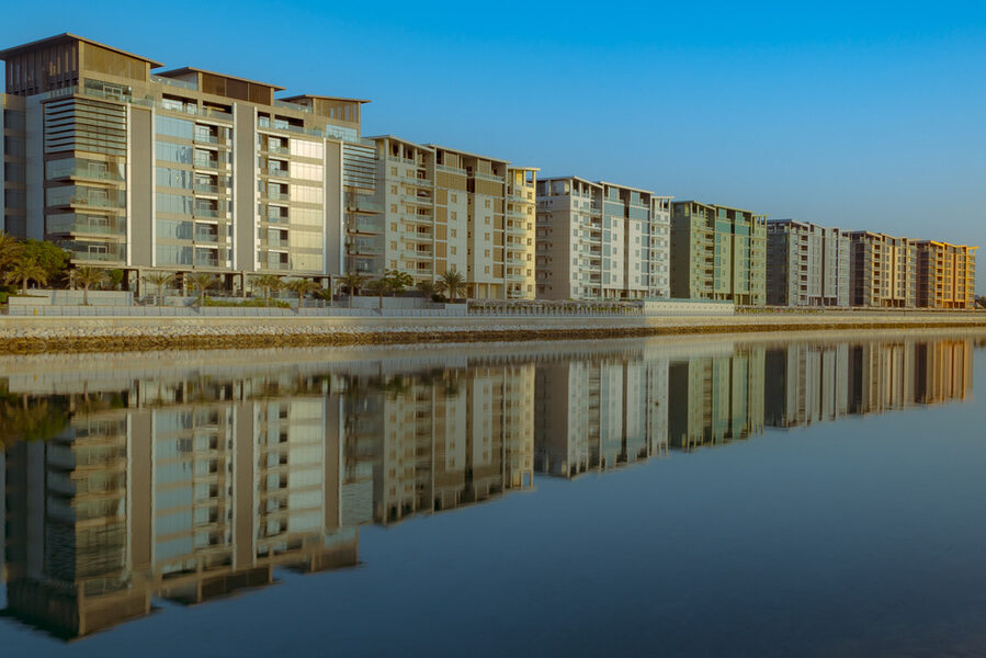 Waterfront apartment buildings reflecting in calm water under a clear sky.