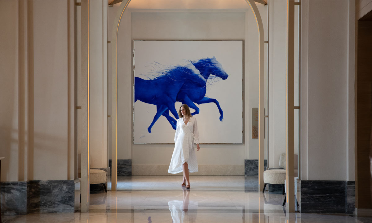 Woman in white dress walking past a painting of blue horses.