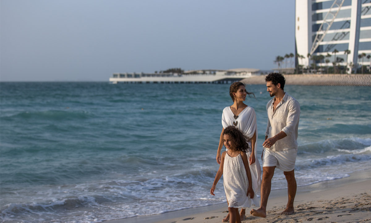 Family walks on a sandy beach near the sea and a modern building.