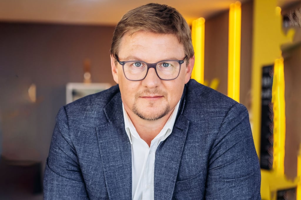 Man in a blazer sitting in a modern space with yellow-lit background.