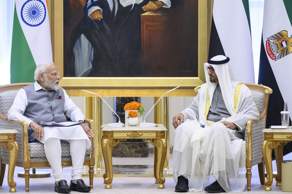 Two men in formal attire converse in opulent room with flags and painting.