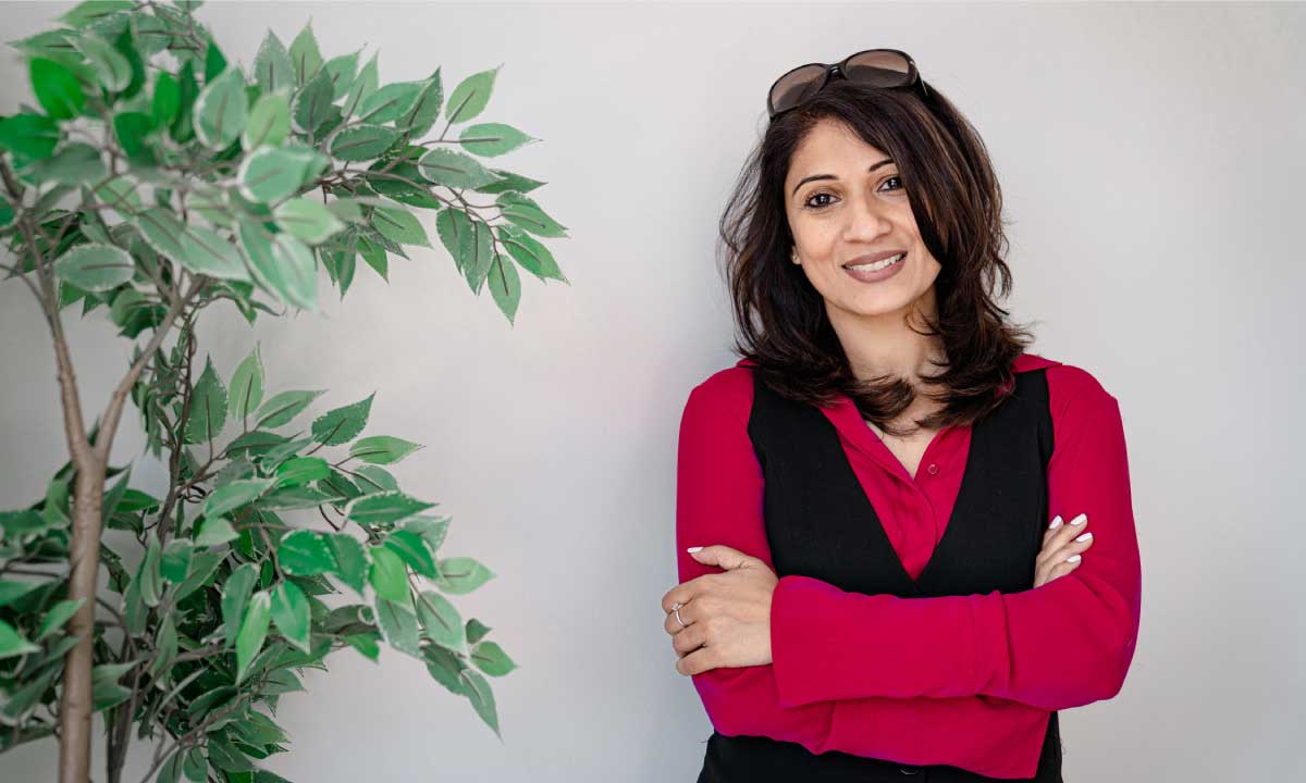 Woman in a red shirt and black vest standing next to a plant.