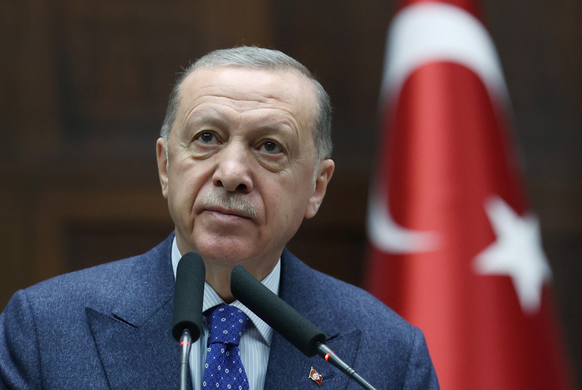 Man in a suit speaking into microphones with Turkish flag in background.