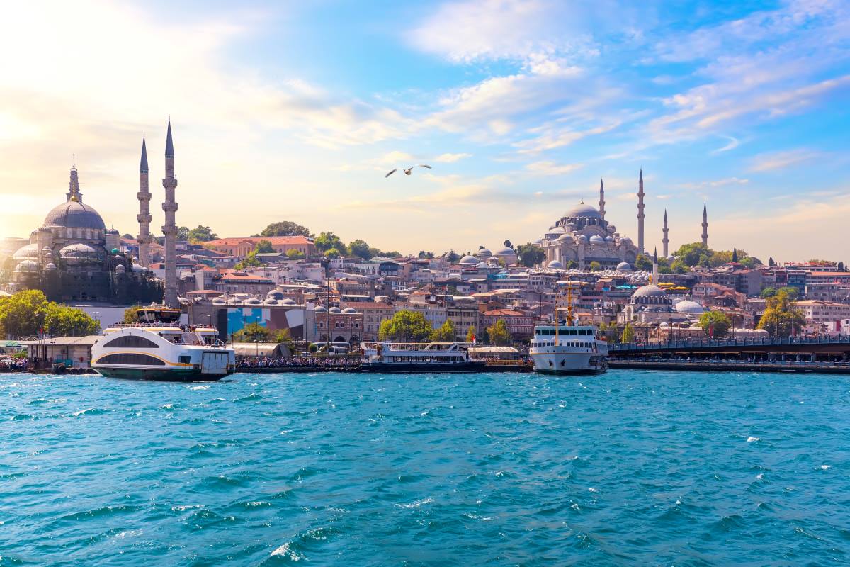 Ferry boats near a waterfront cityscape with mosques under a cloudy sky.