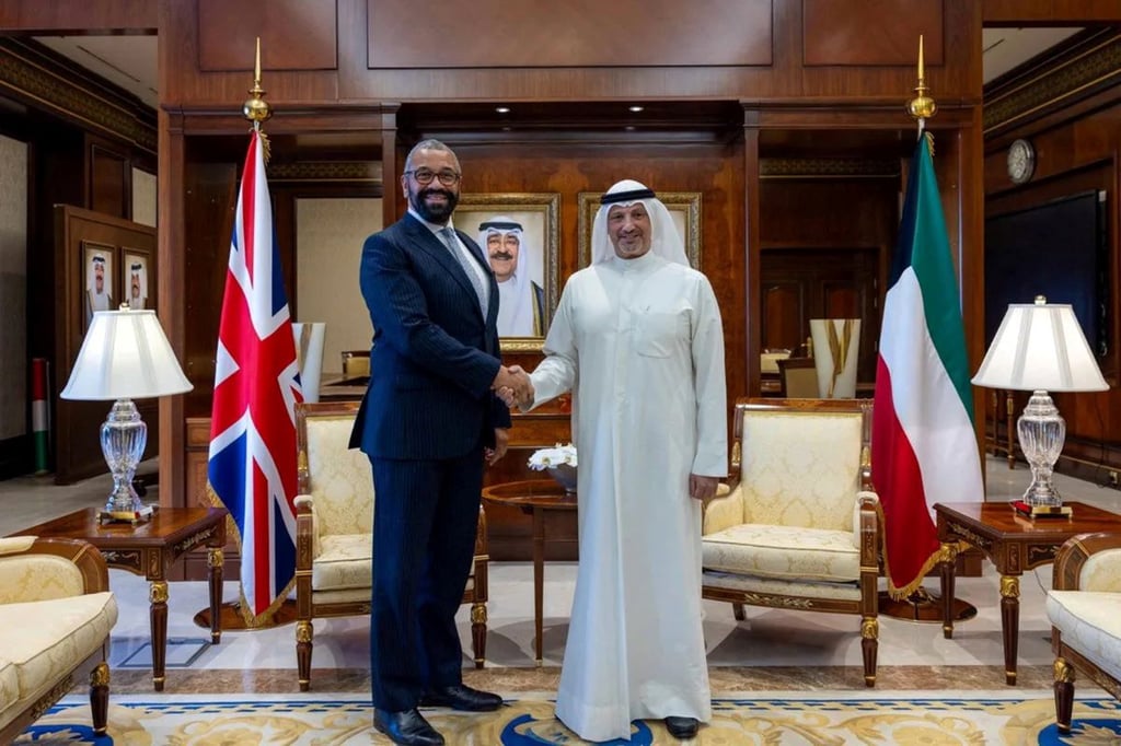 Two men shaking hands, one in suit, other in thawb with flags in background.