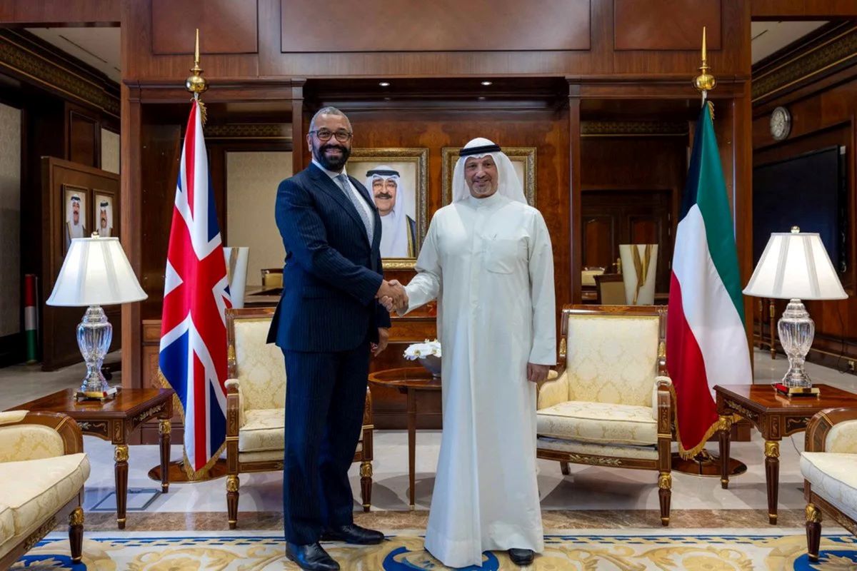 Two men shaking hands, one in suit, other in thawb with flags in background.