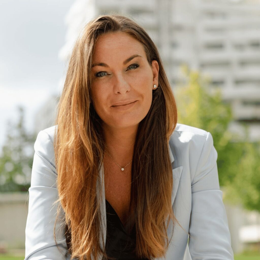 Woman in light blue blazer sits outdoors with blurred background.