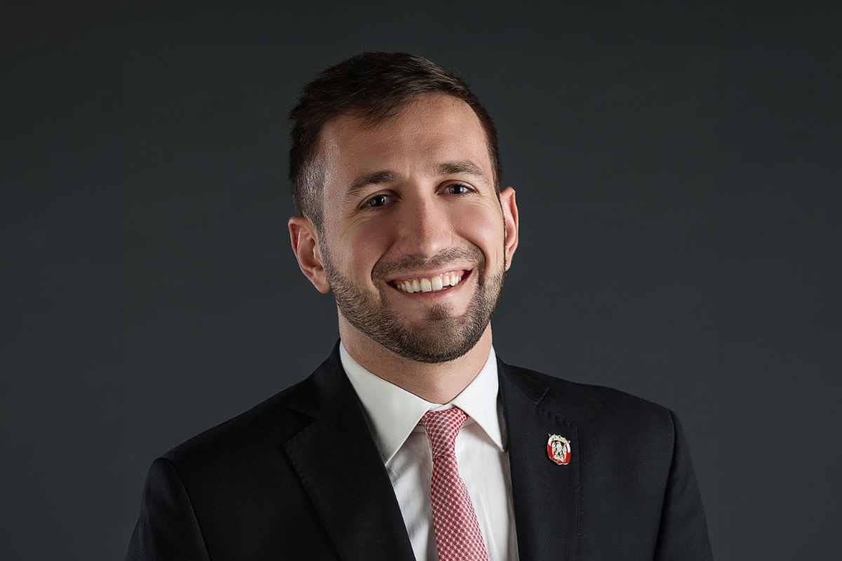 Man smiling in a dark suit with a red tie against a grey background.