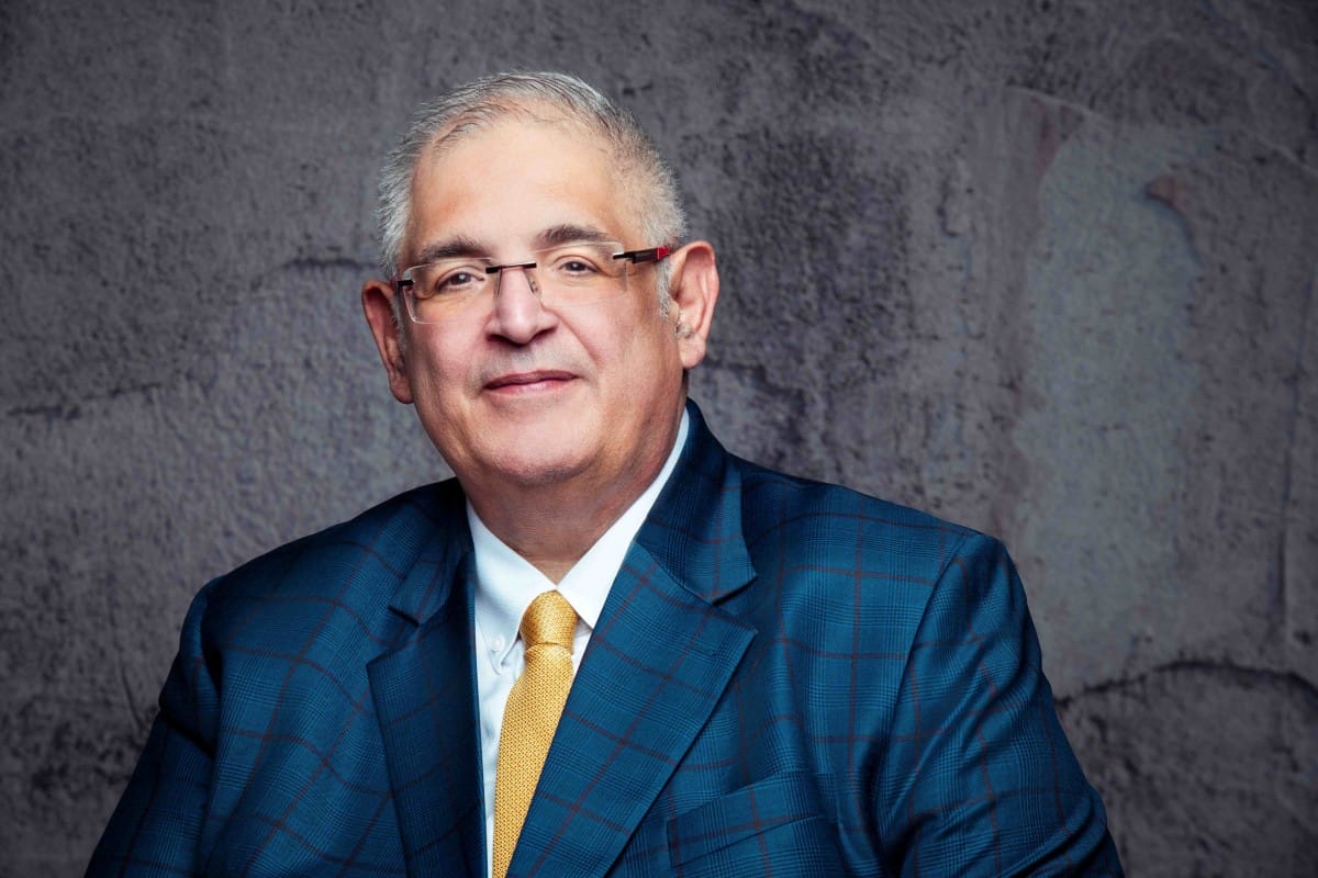 Man in blue checked suit smiling against a gray wall.