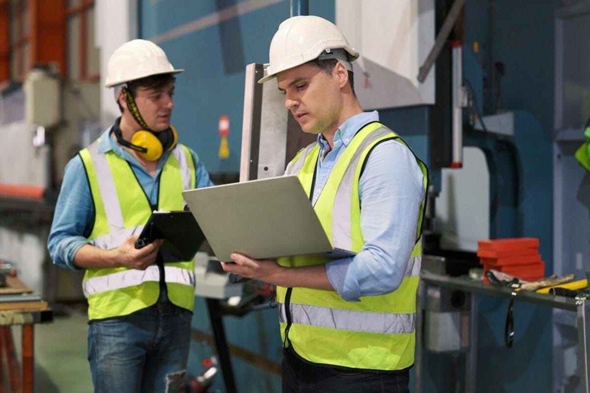 Two men in safety gear reviewing data in a factory setting.