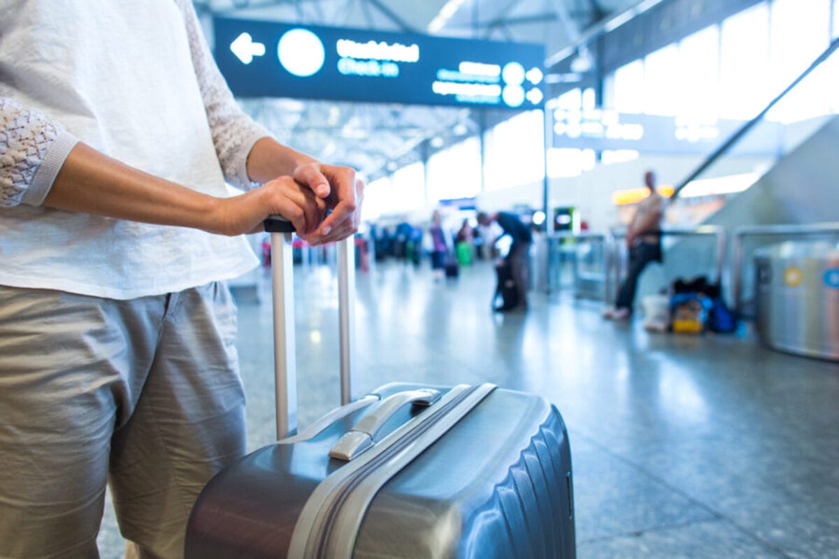 A woman holds a suitcase handle in a busy airport terminal with overhead signs.